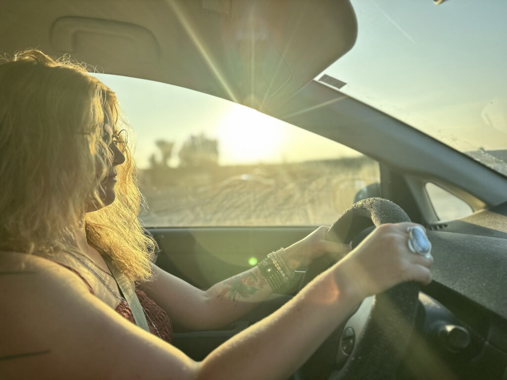 A woman driving a car with the sun set shining through the driver's window illuminating her.