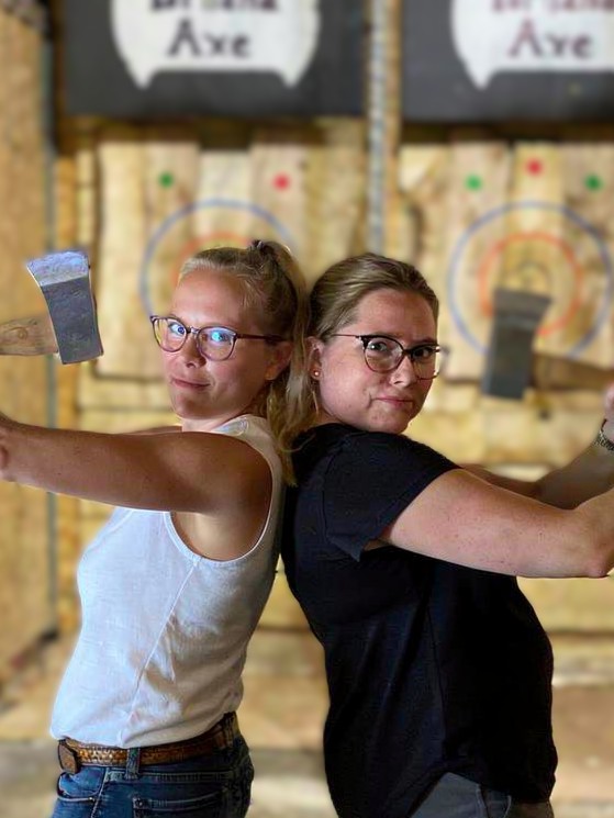 Sue and her sister are standing back-to-back, holding axes at an ax-throwing event—photo taken and copyrighted by Sue Evergreen.