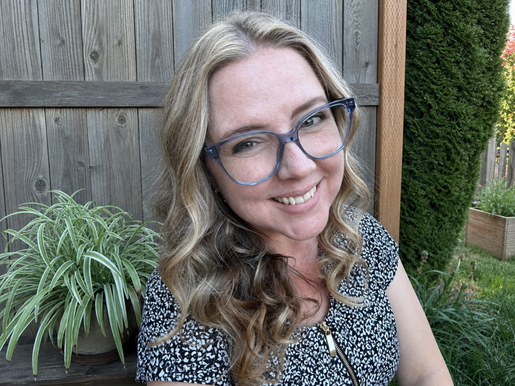 Photo of Kim with glasses, curled haird, and a black and white blouse in her garden.