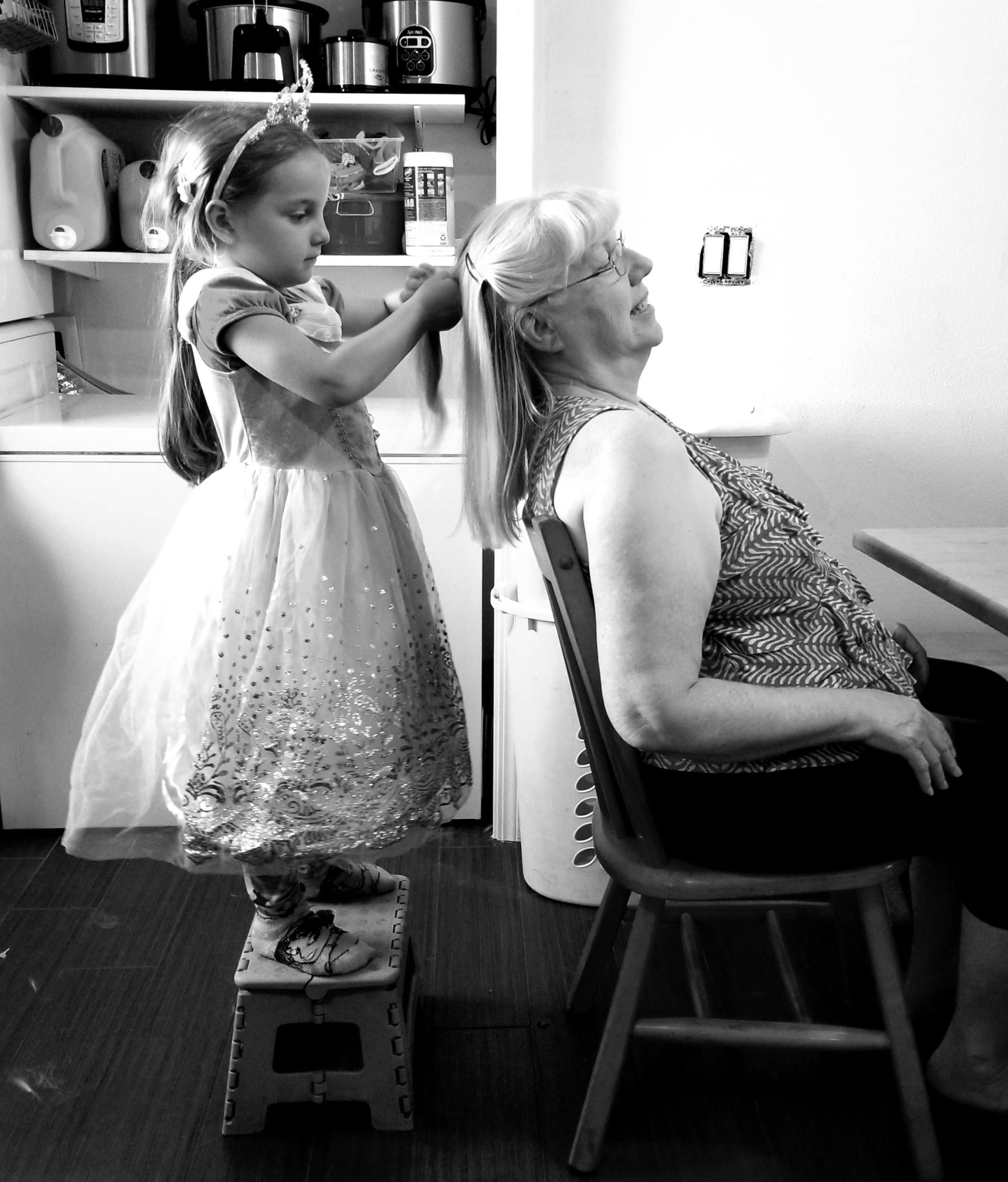 A black-and-white photograph of Sandra, in a chair, with her young granddaughter standing on a stool behind her, doing her hair. A picture of her as a generational matriarch. Photo taken and copyrighted by Sue Evergreen.