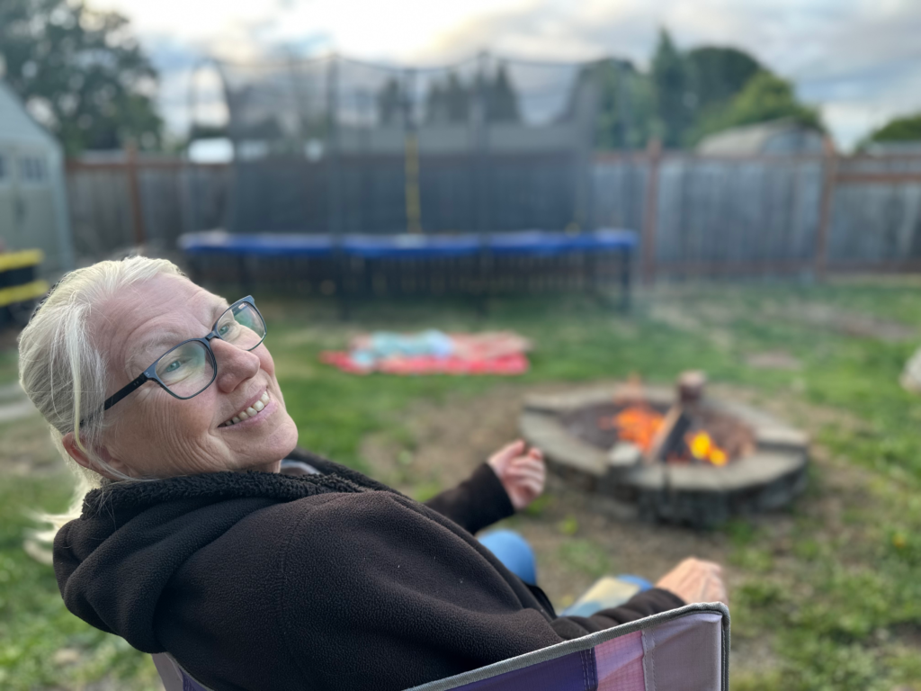 A photo of Sandra. our family matriarch, in 2026 sitting in a lawn chair around a family campfire. Image taken and copyrighted by Sue Evergreen.