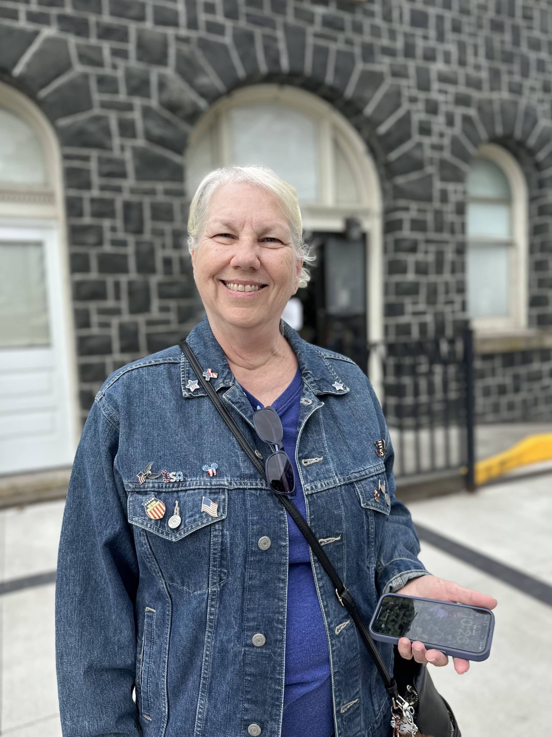 Sandra, now in a jean jacket, is standing in front of a stone building. Photo taken and copyrighted by Sue Evergreen. Sandra's story in "The Matriarchs" Collection.