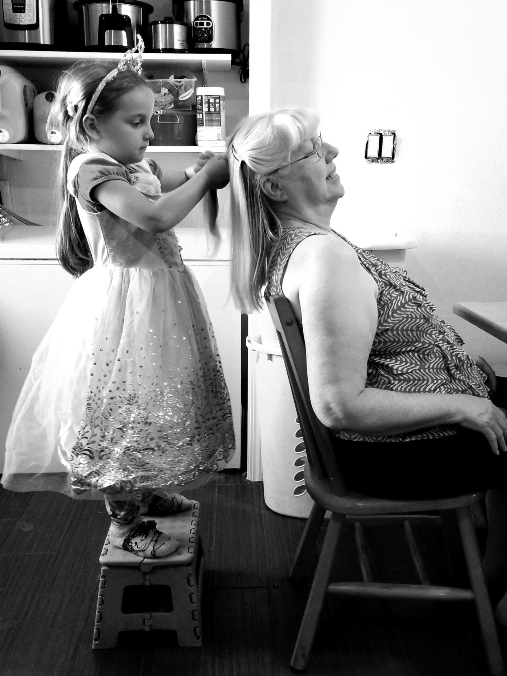 A black-and-white photograph of Sandy in a chair, with her young granddaughter standing on a stool behind her, doing her hair. Copyrighted by Sue Evergreen.