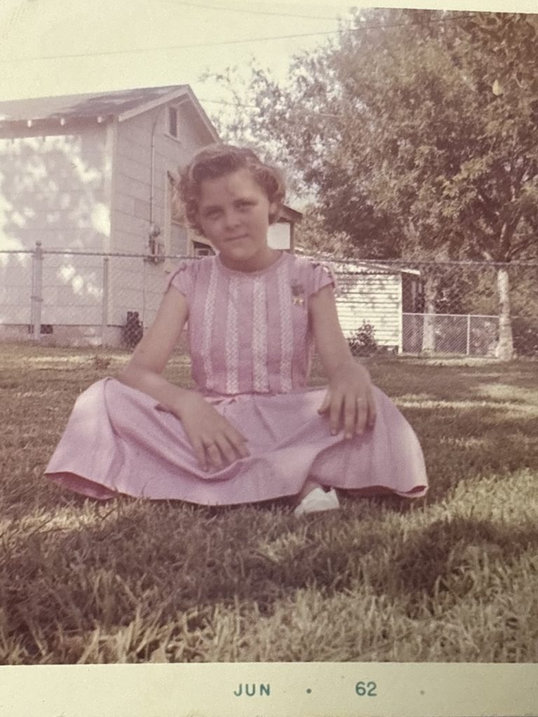 A photo of Suzanne sitting in the grass in a pink dress at age 12 in 1962. Photo permission given to Onward Sister by Suzanne Wright.