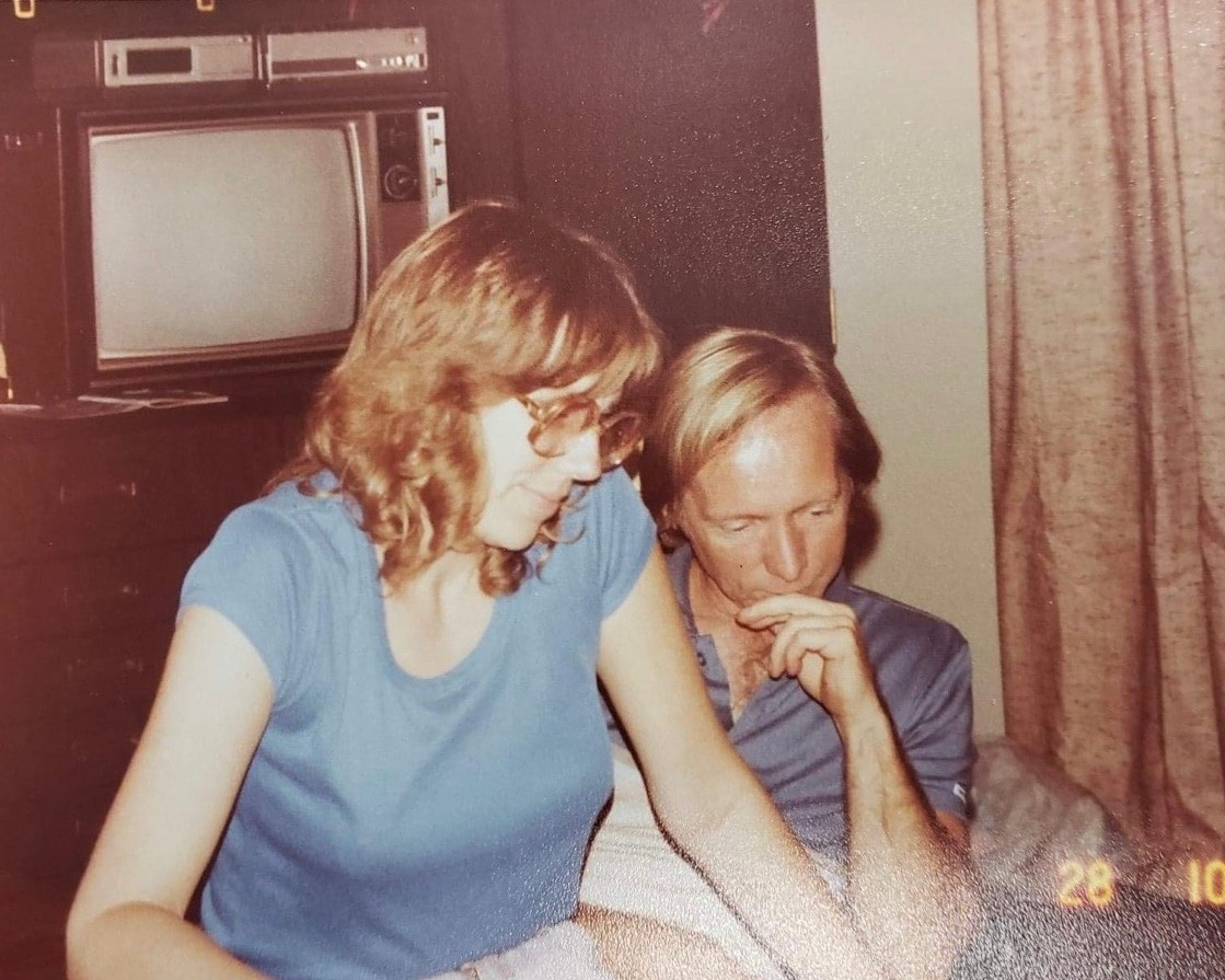 Photo of Sandra and her husband looking at something on a table with a TV behind them in 1976. Photo permissions from Sandra to Onward Sister.