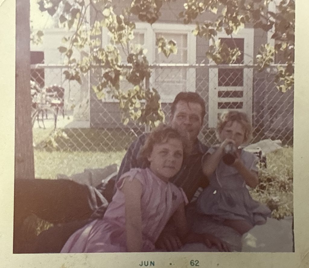 A photo of Suzanne sitting in the grass in a pink dress at age 12 with her father and sister in 1962. Photo permission given to Onward Sister by Suzanne Wright.