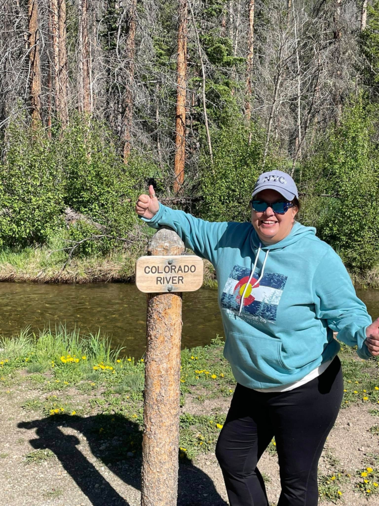 A photo of Natasha in a blue Colorado sweatshirt, hat, and sunglasses next to the Colorado River while hiking for her 50th birthday. Photo permission given to Onward Sister by Natasha Parvey.