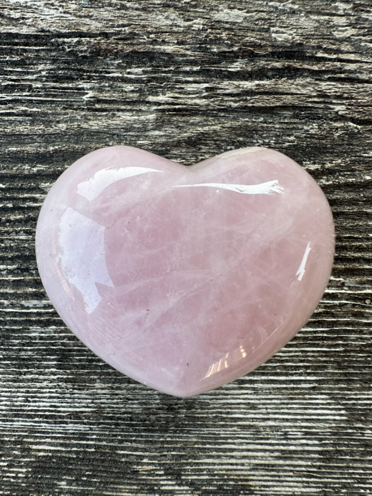 A pink quartz stone heart sitting on a fence board. The stone was given to Sue by her sister during a time of grief. Copyright Sue Evergreen.