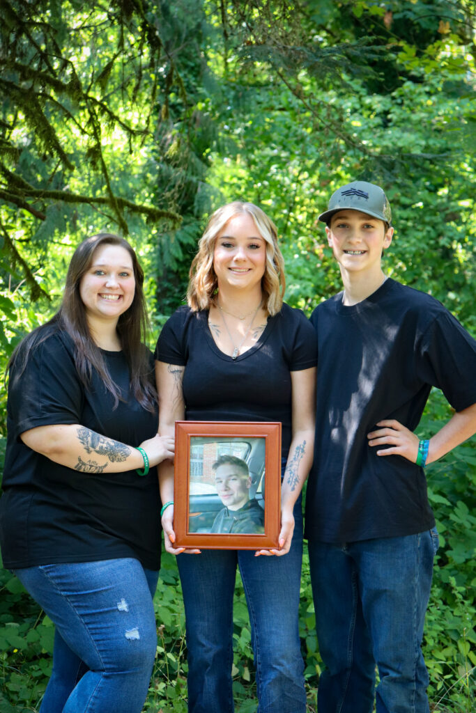 A photo of Sandi's children, from left to right: Megan, Faith, and Nathan. Faith is holding a framed photo of Alex. Photo copyright: Sandi Mueller. Permission for use given to Onward Sister by Sandi Mueller.