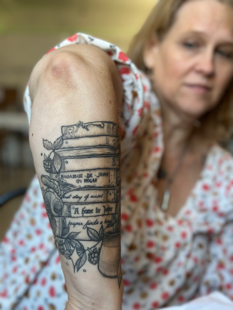 Sandi shows her tattooed forearm of a stack of books. The books are labeled with the children's books she has authored. Photo taken and copyrighted by Sue Evergreen. Permission for use given to Onward Sister by Sandi Mueller.